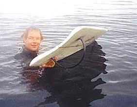 Surfer in a wetsuit floats beside his board, showing its hoop fin