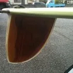 Wide wooden D fin under the tail of a green longboard in a parking lot